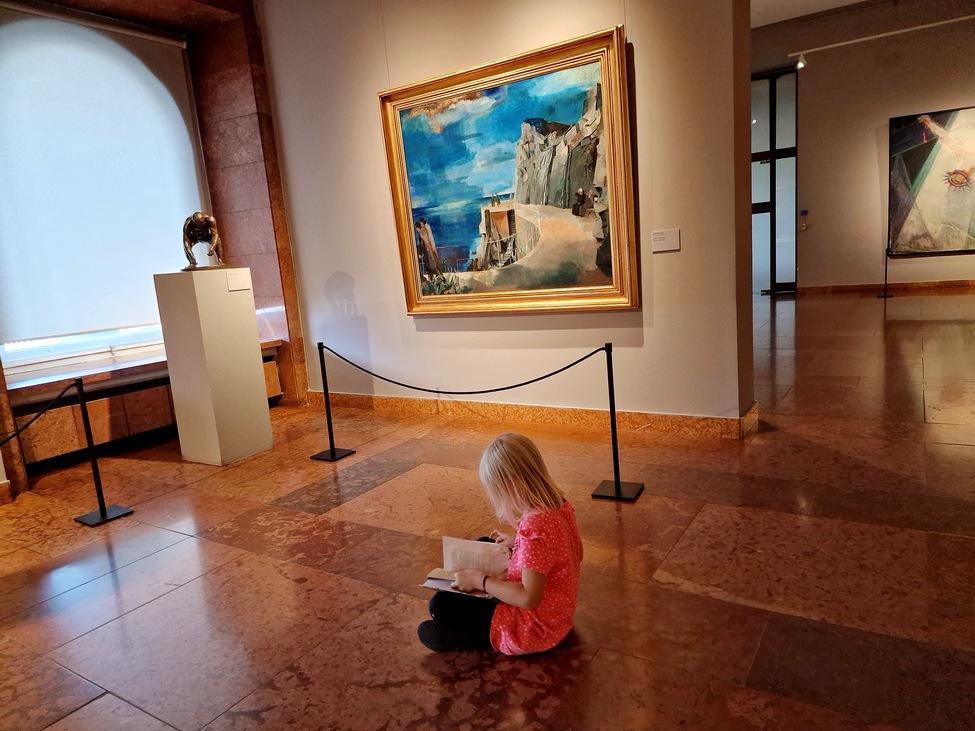 kislány ül a földön könyvet lapozgatva Bernáth Aurél Riviéra című képe előtt